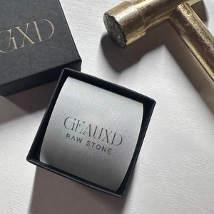 May include: A black box with the text "GXD" on the lid. A smaller black box with a white interior and the text "GEAUXID RAW STONE" is open and sitting on a white surface. A gold hammer is lying on the surface.