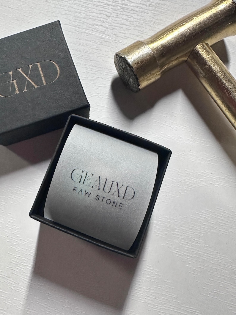 May include: A black box with the text "GXD" on the lid. A smaller black box with a white interior and the text "GEAUXID RAW STONE" is open and sitting on a white surface. A gold hammer is lying on the surface.