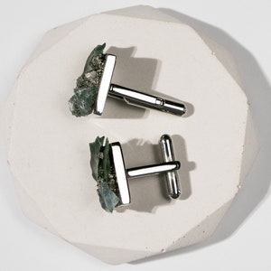 May include: A pair of silver cufflinks with green and gold stone accents. The cufflinks are on a white, textured surface.