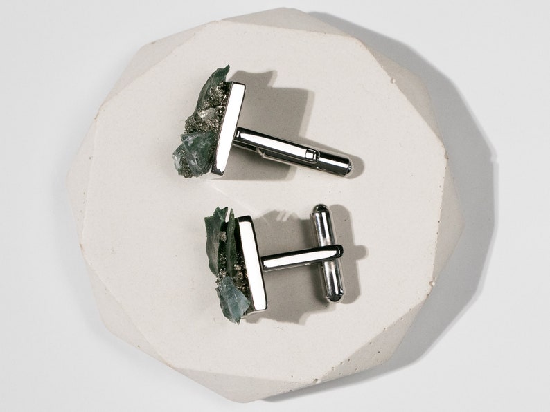 May include: A pair of silver cufflinks with green and gold stone accents. The cufflinks are on a white, textured surface.