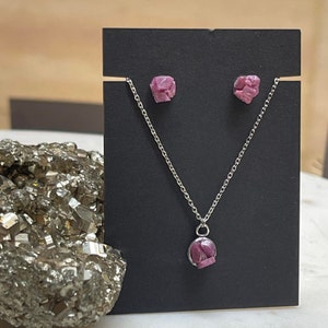May include: A silver necklace and earrings set with pink ruby gemstone pendants. The necklace features a round pendant with a pink ruby gemstone, while the earrings feature square-shaped pink ruby gemstones.