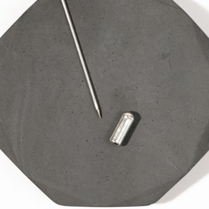 May include: A silver metal pin with a black octagonal base. The pin is lying on a gray surface.