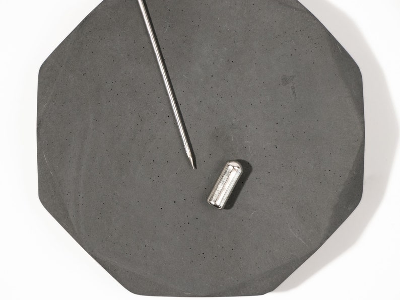 May include: A silver metal pin with a black octagonal base. The pin is lying on a gray surface.