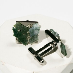 May include: A pair of silver cufflinks with green and blue gemstone accents. The cufflinks are square shaped and feature a cluster of small, irregular shaped stones.