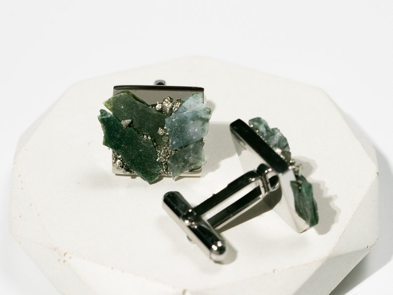 May include: A pair of silver cufflinks with green and blue gemstone accents. The cufflinks are square shaped and feature a cluster of small, irregular shaped stones.