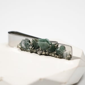 Op de afbeelding: Een zilveren stropdasclip met een cluster van groene en grijze stenen, gezet in een zilveren metalen basis.