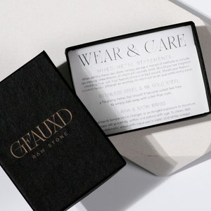 May include: A black box with the words "GEAUXD RAW STONE" in gold lettering. The box is open and a white card with the words "WEAR & CARE" in black lettering is visible. The card contains information about mixed metal statements, stainless steel, 18k gold steel, and raw & satin brass.