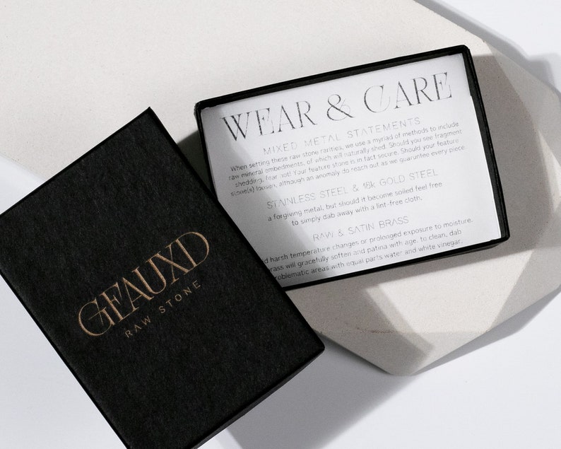 May include: A black box with the words "GEAUXD RAW STONE" in gold lettering. The box is open and a white card with the words "WEAR & CARE" in black lettering is visible. The card contains information about mixed metal statements, stainless steel, 18k gold steel, and raw & satin brass.