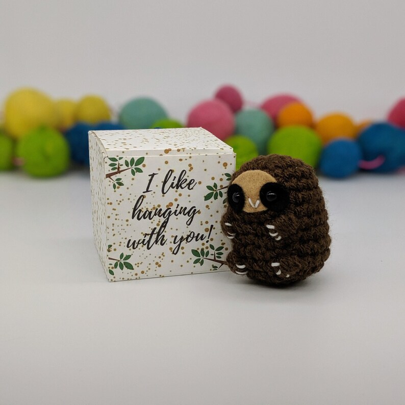 May include: A small brown crocheted sloth with black eyes and a white mouth. The sloth is sitting on a white box with gold glitter and green leaves. The box has the text "I like hanging with you!" printed on it.