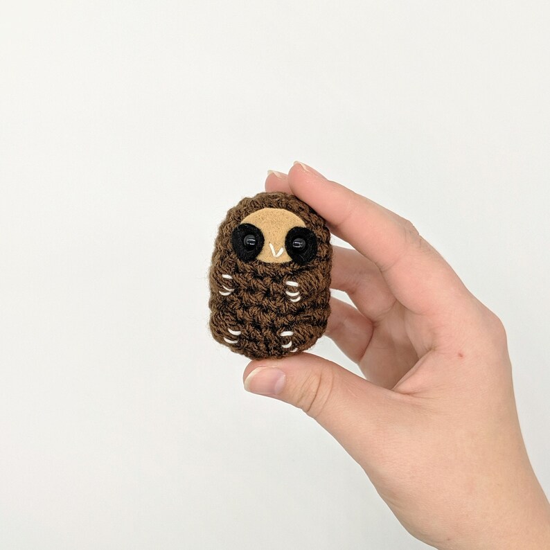 May include: A small brown crocheted owl with black eyes. The owl is curled up in a ball, as if it is sleeping.