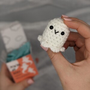 May include: A small, white crocheted ghost with black button eyes and a stitched mouth. The handmade ghost has a rounded body and a ruffled bottom edge. The ghost is held in a person's hand, with a blurred box in the background.