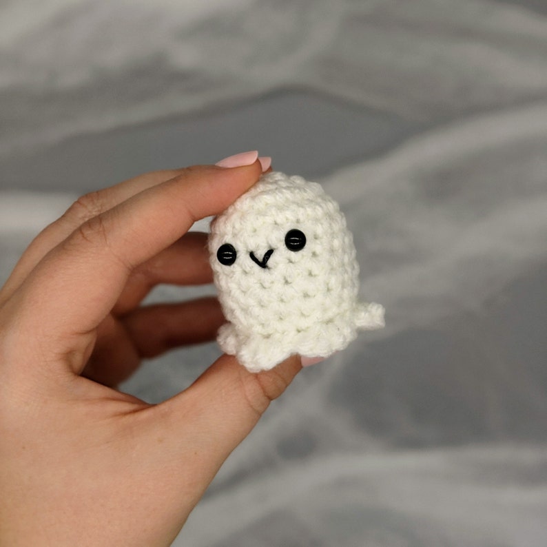 May include: A small, white crocheted ghost with black button eyes and a stitched smile. The handmade amigurumi ghost has a simple, rounded shape with a small, ruffled bottom. The ghost is held in a hand against a soft, gray background.
