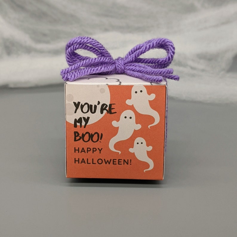 May include: A square Halloween gift box with a purple yarn bow on top. The box is orange with white ghost illustrations and the text "YOU'RE MY BOO! HAPPY HALLOWEEN!" The background is a gray surface.