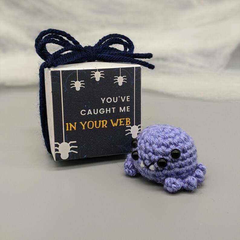 May include: A small, crocheted lavender spider with black eyes and white fangs sits next to a gift box. The box is decorated with spider silhouettes and the text "YOU'VE CAUGHT ME IN YOUR WEB". A navy blue bow secures the box.