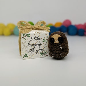 May include: A small brown crocheted sloth with black eyes and a white belly, sitting next to a white gift box with a brown bow and the text "I like hanging with you!" printed on it.