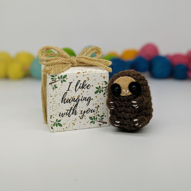 May include: A small brown crocheted sloth with black eyes and a white belly, sitting next to a white gift box with a brown bow and the text "I like hanging with you!" printed on it.
