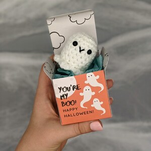 May include: A handmade white crochet ghost with black button eyes and a stitched mouth, presented in a small orange box. The box is decorated with white ghost illustrations and the text "YOU'RE MY BOO! HAPPY HALLOWEEN!"