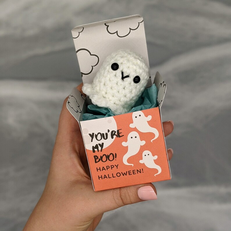 May include: A handmade white crochet ghost with black button eyes and a stitched mouth, presented in a small orange box. The box is decorated with white ghost illustrations and the text "YOU'RE MY BOO! HAPPY HALLOWEEN!"