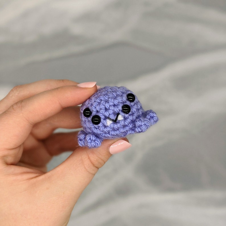 May include: A small, handmade crochet creature in a soft lavender color. It has four black button eyes and a small, smiling mouth with two white teeth. The creature's body is round with small, rounded appendages.