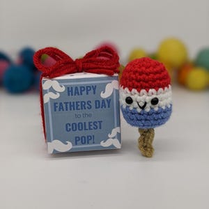 May include: A crocheted popsicle with a smiling face, in red, white, and blue, next to a gift box tied with a red bow. The box has text that reads "HAPPY FATHERS DAY to the COOLEST POP!"