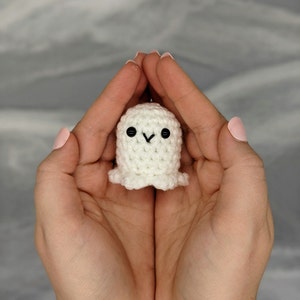 May include: A small, handmade, white crochet ghost with black button eyes and a stitched mouth. The ghost has a rounded body and a ruffled bottom edge. The ghost is held in cupped hands against a soft, blurred background.