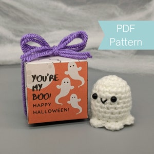 May include: A small white crocheted ghost with black eyes and a smiling mouth. The ghost is sitting next to a small gift box with a purple ribbon. The box has a white background with orange accents and the text "You're my boo! Happy Halloween!" printed on it.