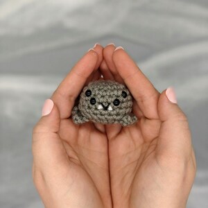 May include: A small, crocheted gray creature with black eyes and white teeth is held in cupped hands. The handmade toy has a rounded body and small, stubby legs. The background is a soft, blurred gray.