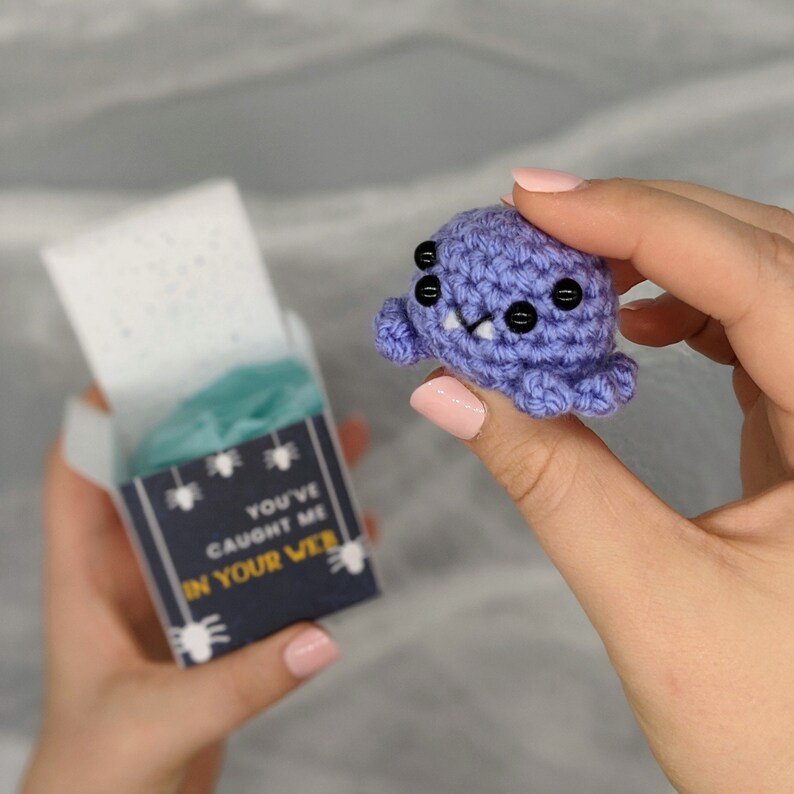 May include: A small, crocheted purple spider with black button eyes and a smiling mouth is held near a small gift box. The box has the text "YOU'VE CAUGHT ME IN YOUR WEB" and spider illustrations. The spider is approximately 1.5 inches in diameter.