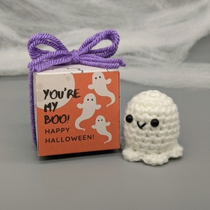 May include: A white crocheted ghost with black eyes and a smiling mouth sits next to a small gift box. The box is orange with white ghost illustrations and the text "YOU'RE MY BOO! HAPPY HALLOWEEN!" A purple yarn bow adorns the box.