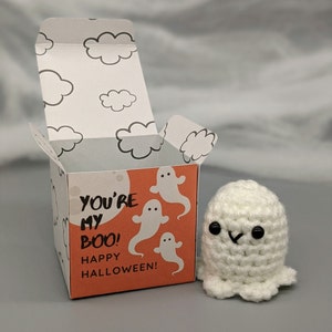 May include: A white crocheted ghost figure next to a Halloween-themed gift box. The box is orange with white ghost illustrations and the text "YOU'RE MY BOO! HAPPY HALLOWEEN!". The box's interior and lid feature cloud designs.