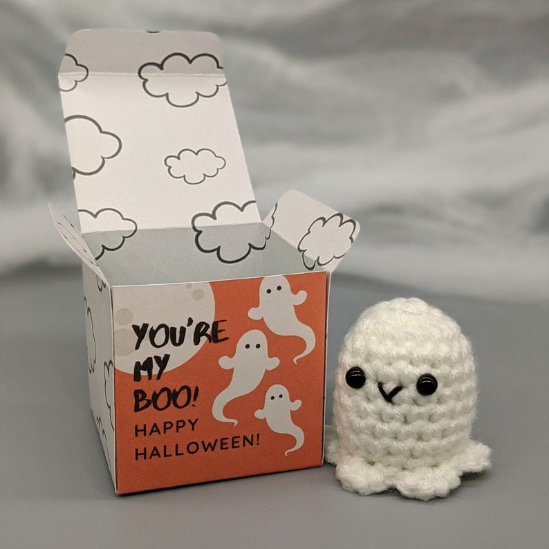 May include: A white crocheted ghost figure next to a Halloween-themed gift box. The box is orange with white ghost illustrations and the text "YOU'RE MY BOO! HAPPY HALLOWEEN!". The box's interior and lid feature cloud designs.
