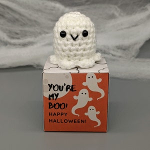May include: A handmade, white crochet ghost with black eyes sits atop a small orange box. The box features the text "YOU'RE MY BOO! HAPPY HALLOWEEN!" and illustrations of white ghosts. The background includes a gray surface and white spiderweb decor.