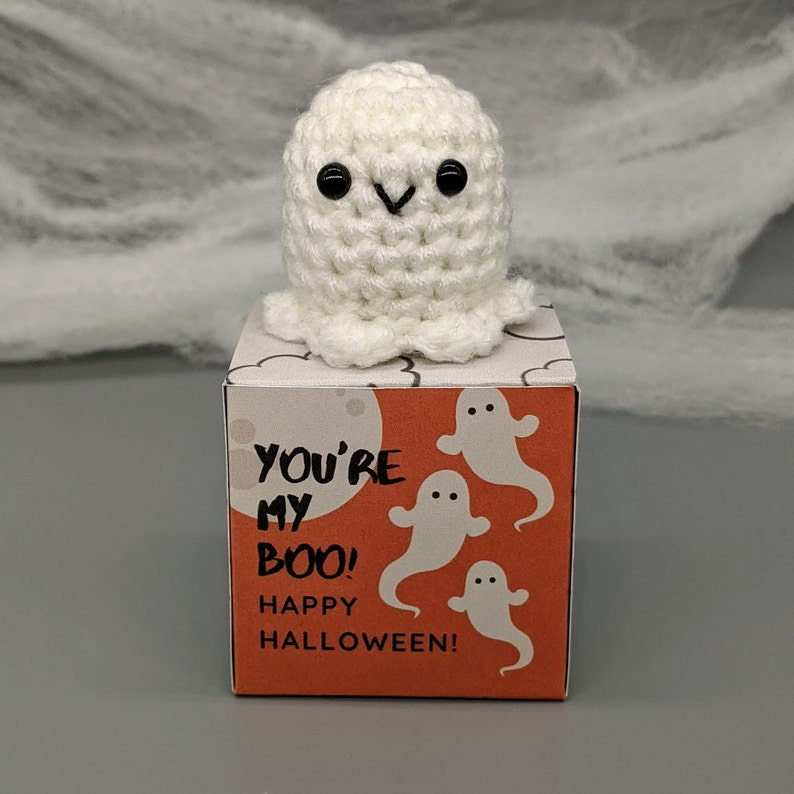 May include: A handmade, white crochet ghost with black eyes sits atop a small orange box. The box features the text "YOU'RE MY BOO! HAPPY HALLOWEEN!" and illustrations of white ghosts. The background includes a gray surface and white spiderweb decor.