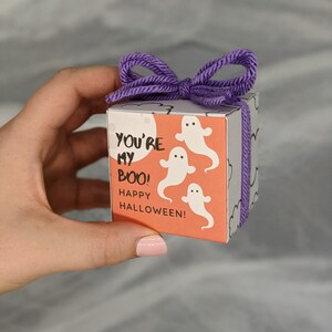 May include: A small, square gift box with a purple yarn bow. The box features a Halloween theme with white ghost illustrations and the text "YOU'RE MY BOO! HAPPY HALLOWEEN!" on an orange background.