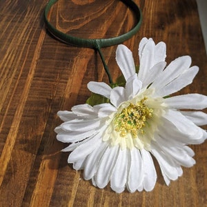 May include: A white daisy flower with a green stem and leaves attached to a green headband.