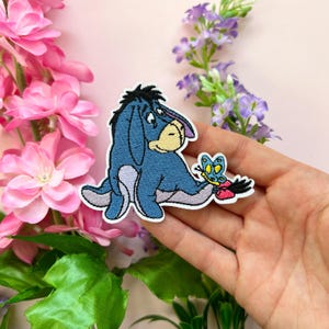 Op de afbeelding: Geborduurde strijkpatch met een blauw ezel-personage en een vlinder. De ezel kijkt naar de vlinder, die roze, geel en blauw is.