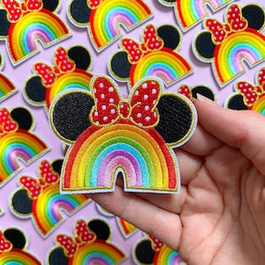 Op de afbeelding: Geborduurde patch met een regenboogboog en een Minnie Mouse-hoofdontwerp. De patch heeft een rode strik met witte stippen, zwarte oren en een gouden rand. De regenboog toont levendige kleuren, waaronder rood, oranje, geel, groen, blauw en paars.