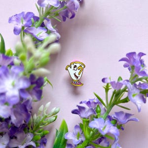May include: A colourful enamel pin of Chip from Beauty and the Beast. The pin shows Chip's smiling face with a pink rim, yellow handle, and a decorative base. The pin is set against a soft pink background, surrounded by purple flowers.