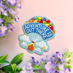 May include: A blue embroidered patch featuring a hot air balloon carrying a house. The balloon is filled with colourful balloons and the patch reads "Adventure is out there".