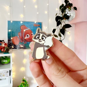 Op de afbeelding: Een emaille pin van een wasbeer in lichtbruine en crèmekleurige tinten, die een wit object vasthoudt. De pin heeft een goudkleurige omtrek. Op de achtergrond een ingelijste afbeelding van een clownvis.