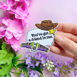 Può includere: Toppa ricamata con cappello da cowboy marrone, stella gialla e design navicella spaziale di Buzz Lightyear. La toppa include il testo "You've got a friend in me" in una nuvoletta. Fiori rosa e viola sullo sfondo.