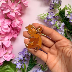 Pode incluir: Um alfinete de esmalte marrom e laranja com um urso e seu filhote. O urso está sentado com o filhote nas costas. O alfinete tem um contorno dourado e está sobre um fundo rosa com flores.