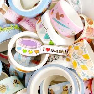 Op de afbeelding: Een gouden emaille pin in de vorm van een rol washi tape met een rood hart en de tekst "Ik hou van washi" erop. De pin is omringd door rollen kleurrijke washi tape.