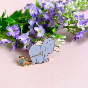 May include: A gold-tone enamel pin depicting a blue elephant being pulled by a small, blue, mouse-like creature. The pin is displayed against a pink background with purple flowers.