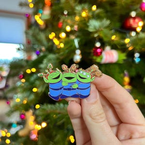Op de afbeelding: Een speld met drie groene alien-personages uit Toy Story, gekleed in blauwe outfits en feestelijke hoofddeksels. De speld wordt vastgehouden tegen een achtergrond van een kerstboom met kleurrijke lichten en versieringen.