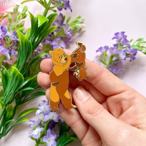 Puede incluir: Un pin de esmalte dorado con dos osos de dibujos animados. Un oso está de pie y el otro está colgado boca abajo. Los osos son de color marrón y beige con contornos negros. El pin se sostiene en una mano sobre un fondo de flores moradas.