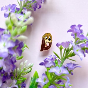 Op de afbeelding: Een goudkleurige emaille pin met een cartoonhond met bruine oren en een roze tong. De hond lacht met zijn tong uit. De pin staat tegen een lichtroze achtergrond met paarse bloemen.
