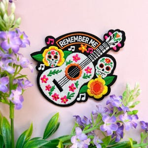 Op de afbeelding: Geborduurde strijkpatch met een witte gitaar met kleurrijke bloemen en twee suiker schedels. De gitaar heeft de tekst "Remember Me" bovenaan.