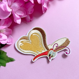Op de afbeelding: Geborduurde vlinderpatch met een hartvormig lichaam in tinten goud, bruin en wit, omlijnd in wit. De vlinder heeft een rode mond en antennes, tegen een roze achtergrond met roze bloemen.