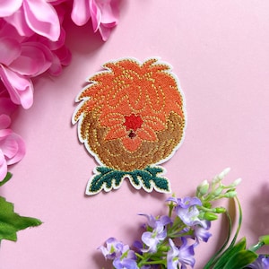 Op de afbeelding: Geborduurde strijk-patch met een gestileerde oranje bloem met een bruin centrum en groene bladeren. De bloem heeft een gouden glitter-effect.